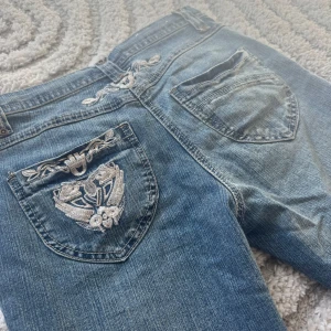 Blå jeans med broderad bakficka från 2000st - Snygga blå jeans med broderad detalj på ena bakfickan. Jeans med dragkedja och knapp. Jeansen är bootcut benlängden är runt 80💕
