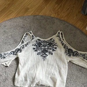 Vit blus med broderi och öppna axlar - Säljer en vit blus med svarta broderade mönster på bröstet och ärmarna. Blusen har öppna axlar och långa ärmar samt dekorativa snören med tofsar vid halsen. Perfekt för en bohemisk stil.