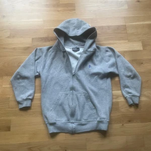 Ralph Lauren Hoodie - Grå hoodie med dragkedja från Ralph Lauren. Klassisk modell med huva, fickor framtill och broderad blå logga på bröstet. Perfekt för en avslappnad stil och enkel att matcha med det mesta. Storleks lappen är borta så vet inte vad storleken är så kolla måtten noggrant men jag skulle tro att den är XS :) har några vit fläckar och andra defekter, skriv för att få bilder då jag inte får plats med dem i annonsen 😊