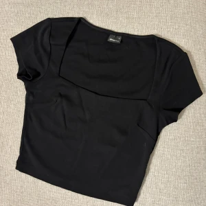 Svart croppad topp från Gina Tricot - Säljer en svart croppad topp från Gina Tricot med korta ärmar och fyrkantig ringning. Perfekt att matcha med höga jeans eller kjol. Enkel och stilren design som passar till många tillfällen.