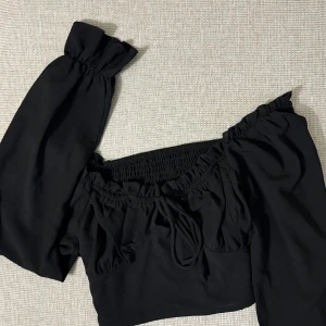Svart offshoulder topp från SHEIN - Säljer en svart offshoulder topp från SHEIN med långa puffärmar och smockad resår vid axlar och midja. Toppen har knytning framtill och en luftig, trendig passform. Perfekt för dig som vill ha en snygg och bekväm topp till garderoben.