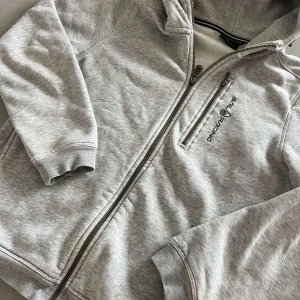Grå hoodie från Sail Racing - Säljer en ljusgrå hoodie från Sail Racing med dragkedja framtill och en bröstficka med dragkedja. Jackan har huva och ribbade muddar vid ärmslut och nederkant. Perfekt för en sportig och avslappnad stil.