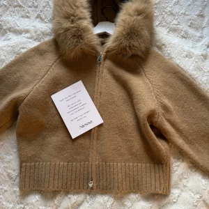Beige ulljacka med pälskantad huva från Alessa - Mysig beige jacka i ull med dragkedja och stor huva kantad med fluffig päls. Jackan har ribbade muddar och är perfekt för kyliga dagar. Snygg och enkel att styla till olika outfits. Helt ny aldrig använd (bara provat den) pris kan sänkas
