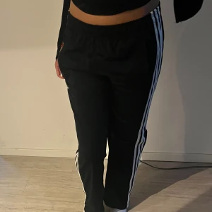 Svarta träningsbyxor från Adidas med vita ränder - Svarta träningsbyxor från Adidas med klassiska vita ränder längs sidorna och Adidas-logga på ena benet. Byxorna har resår i midjan och är perfekta för sport eller chill. Snygg och enkel design som funkar till mycket.