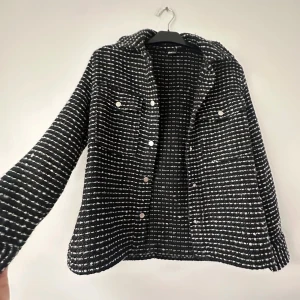 Svartvit rutig jacka med knappar - Säljer en svartvit jacka med rutigt mönster och silvriga knappar framtill. Jackan har lång ärm och en klassisk krage. Perfekt för dig som gillar en stilren och trendig look.