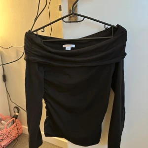 Svart offshoulder topp - Säljer en svart offshoulder topp med långa ärmar. Toppen har en bred krage som vilar över axlarna och är figurnära i modellen. Perfekt för en trendig och stilren look.