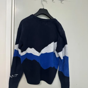 Mörkblå stickad tröja med bergsmotiv från GANT - Säljer en mörkblå stickad tröja från GANT med motiv av berg och linbana i vitt och blått. Tröjan har rund halsringning och långa ärmar. Perfekt för dig som gillar unik design och varma plagg.