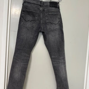 Grå jeansbyxor med raka ben - Säljer ett par grå jeansbyxor med klassisk femficksdesign och raka ben. Jeansen har svarta detaljer vid midjan och subtila slitningar för en snygg, använd look. Passar perfekt till vardags och är lätta att matcha med olika stilar.