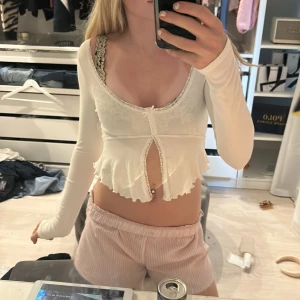 Vit croppad topp med volang från Gina Tricot - Säljer en söt vit croppad topp från Gina Tricot med volangkant och dekorativ spets längs ringningen. Toppen har långa ärmar och knyt framtill. Perfekt för att skapa en romantisk och trendig look.