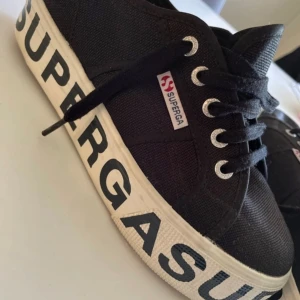 Svarta sneakers från Superga med stor logga - Säljer ett par svarta Superga-sneakers i tyg med kraftig vit sula och stor svart logga runt hela sulan. Skorna har snörning och silverfärgade öljetter. Perfekta för dig som gillar en sportig och trendig stil. Använda ett par gånger. Köpt i Italien för ca 1000kr. 