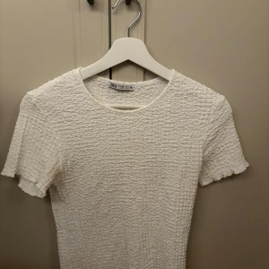 Vit ribbad topp från Zara - Säljer en vit ribbad topp från Zara med korta ärmar och rund halsringning. Toppen har en strukturerad, stretchig yta och sitter figurnära. Inga defekter. Perfekt till många olika outfits!