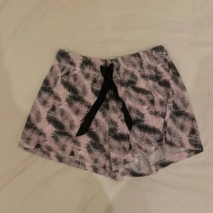 Rosa och svarta shorts med palmbladsmönster - Säljer ett par rosa pyjamas shorts med svart palmbladsmönster och svart knytband i midjan. Shortsen är tillverkade i en mjuk blandning av bomull och polyester, perfekta för varma dagar.