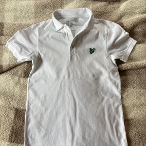 Vit pikétröja från Lyle & Scott - Klassisk vit pikétröja från Lyle & Scott med korta ärmar och krage. Dammodell. Har en knappt syrlig fläck (se sista bilden) därav priset. 