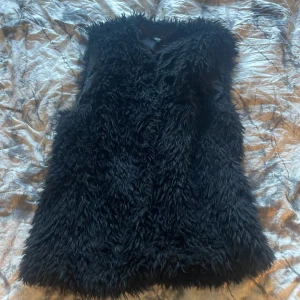 Svart pälsväst - Säljer en svart pälsväst med fluffig och mjuk känsla. Perfekt att slänga över en hoodie eller tröja för en cool look. Västen har en rak passform och är helsvart utan synliga detaljer framtill.