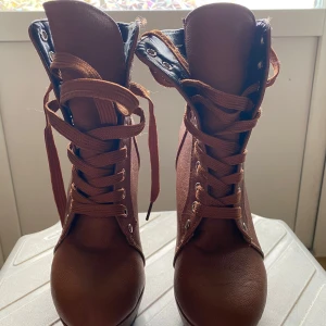 Bruna höga boots med snörning och klack - Säljer ett par bruna boots med hög klack och platåsula. Skorna har snörning framtill, två dekorativa spännen på sidan och rund tå. Tillverkade i syntetmaterial och passar perfekt för dig som vill sticka ut.