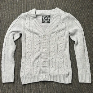 Ljusgrå stickad kofta med knappar - Ljusgrå stickad kofta med kabelstickat mönster och fyra stora knappar framtill. V-ringning och ribbade muddar vid ärmslut och nederkant. Perfekt för lager-på-lager och enkel att matcha. Storlek M. Hör av er vid frågor och funderingar!