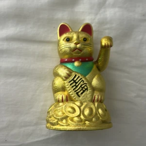 Maneki-neko lyckokatt i guld - En klassisk Maneki-neko lyckokatt i guld med röd krage och turkos detalj. Katten sitter på en hög av guldmynt och håller upp ena tassen. Perfekt som dekoration eller lyckobringare.