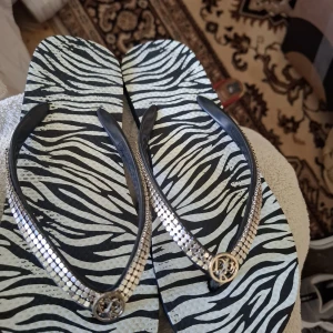 Zebramönstrade sandaler med silverdetaljer - Säljer ett par sandaler med svartvitt zebramönster och remmar i svart med silvriga detaljer och ett runt emblem framtill. Perfekta för sommaren och ger en cool touch till din outfit.