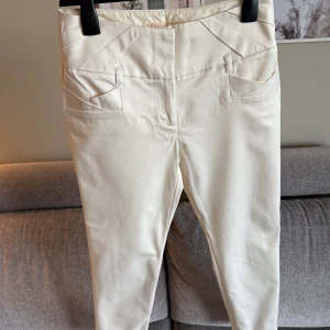 Vita kostymbyxor med hög midja - •Eleganta, benvita byxor från Orsay.  •Byxorna har en unik design med dekorativa detaljer vid fickorna och en smickrande passform. •Har raka ben, diskreta fickor både fram och bak samt stängs med hyska och hake. •Perfekt för en stilren och sofistikerad look. •Djurfritt och rökfritt hem.