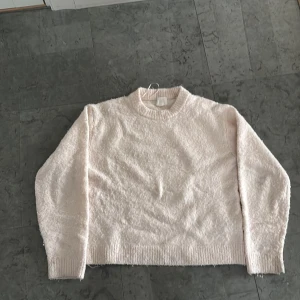 Ljusrosa fluffig stickad tröja - Mysig ljusrosa stickad tröja med rund halsringning och långa ärmar. Tröjan har en fluffig textur och ribbade muddar vid ärmslut och nederkant. Perfekt för kyligare dagar.
