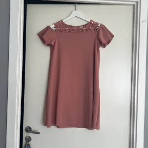 Rosa klänning med spetsdetalj - Söt rosa klänning från Lindex med volangärmar och spets upptill. Klänningen har offshoulder-design. Mjuk och skön i materialet, lätt att bära hela dagen. I princip oanvänd!