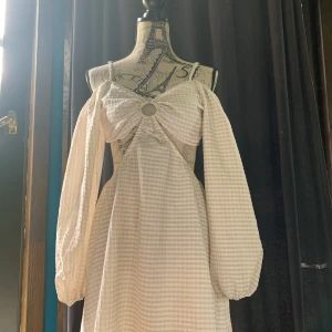 Beige rutig kortklänning - Supersöt kortklänning i beige och vitt rutigt från H&M Divided. Klänningen har långa puffärmar, offshoulder-design och en snygg ringdetalj framtill. Tillverkad i luftig bomull, perfekt för varma dagar. Smockad rygg för extra komfort och snygg passform. Knappt använd. 