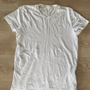 Vit t-shirt från GANT i bomull - Klassisk vit t-shirt från GANT i mjuk bomull. T-shirten har rund halsringning, korta ärmar och en liten GANT-logga tryckt på bröstet. Enkel och stilren modell som passar till allt. Perfekt basplagg för garderoben.