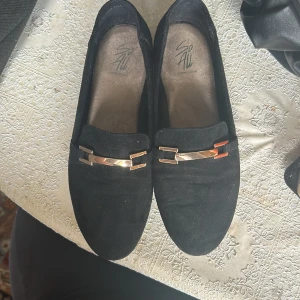 Svarta mockaloafers med gulddetalj - Stilrena svarta loafers i mocka från So. Skorna har platt sula och rund tå. .