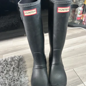 Svarta höga stövlar från Hunter - Klassiska svarta höga stövlar från Hunter. Tillverkade i slitstarkt gummi, perfekta för regniga dagar. Snygg logga framtill och tidlös design som funkar till många outfits.