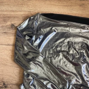 Glittrig silver långärmad topp - Snygg långärmad topp i glittrigt silvermaterial med figurnära passform. Perfekt för dig som vill sticka ut och glänsa. Toppens material är syntet och den har en cool metallic-look som drar blickarna till sig.