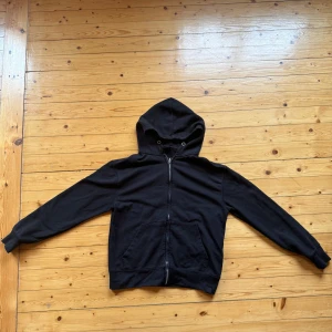 Svart hoodie från Pull&Bear med dragkedja - Svart hoodie från Pull&Bear i storlek M. Klassisk modell med huva, snörning och hel dragkedja framtill. Mjukt bomullsmaterial och två fickor framtill. Perfekt för en chill och avslappnad stil.