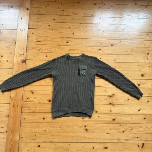 Olivgrön stickad tröja Lyle & Scott - Säljer en olivgrön stickad tröja från Lyle & Scott med ribbad struktur och en snygg bröstficka med lock och gul logga. Tröjan har rund hals och långa ärmar, perfekt för lager-på-lager under kyliga dagar.