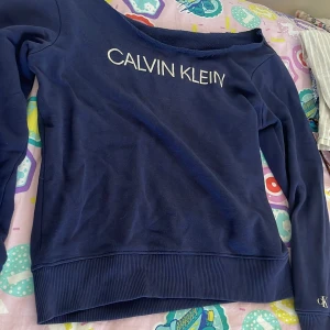 Mörkblå Calvin Klein sweatshirt - Mörkblå sweatshirt från Calvin Klein med vit logga framtill och 'JEANS'-patch baktill. Tröjan har bred ribbad mudd i nederkant och vid ärmslut, samt lång ärm. Skön och avslappnad passform, perfekt till jeans eller joggers.