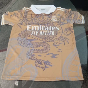 Real Madrid Ronaldo draktröja XS - Unik Real Madrid fotbollströja i guldbeige med stort drakmotiv och vita detaljer. Nummer 7 och Ronaldo på ryggen, klassisk vit krage och klubbmärke på bröstet. Tillhörande vita shorts med guldiga ränder och nummer 7. Perfekt för fans av Ronaldo.