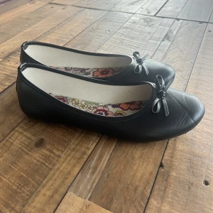Svarta ballerinaskor med rosett - Snygga svarta ballerinaskor med rund tå och liten rosett framtill. Skorna har en blommig innersula som ger en extra touch. Perfekta för dig som gillar en klassisk och söt stil. Tillverkade i skinnliknande material.