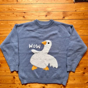 Blå stickad tröja med gås från Aelfric Eden - Superchill blå stickad tröja från Aelfric Eden med en stor fluffig gås och texten 'WOW' på framsidan. Tröjan har rund hals, långa ärmar och ribbade muddar. Perfekt för dig som gillar unika och roliga plagg!
