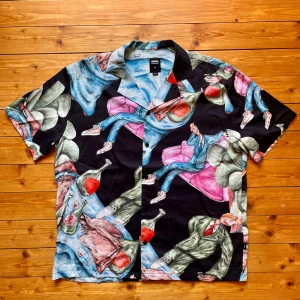 Färgstark skjorta från Dr Denim M - Unik skjorta från Dr Denim i storlek M med färgstarkt konstnärligt mönster som föreställer personer, flaskor och soffor i blått, rosa, grönt och rött mot svart bakgrund. Skjortan har korta ärmar, knappar framtill och en avslappnad passform, oversized. Perfekt för dig som vill sticka ut.