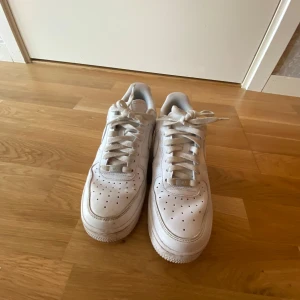 Vita Nike Air Force 1 sneakers - Klassiska vita Nike Air Force 1 sneakers med lågt skaft och perforerad tå för extra ventilation. Skorna har snörning, rund tå och är tillverkade i skinn med en platt sula. Perfekta för en clean och stilren look.