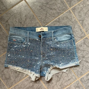 Jeansshorts med paljetter från Hollister - Blå jeansshorts från Hollister med snygga paljettdetaljer framtill och råa, fransiga kanter. Klassisk femficksmodell, låg midja och normal passform. Perfekta för sommaren när du vill sticka ut lite extra.