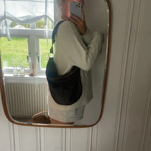 Brun axelväska i skinn med dragkedja - Säljer en brun axelväska i mjukt skinn med snygga mässingsfärgade ringar och dragkedja upptill. Väskan har en rundad form och bred axelrem, perfekt för att bära över axeln. Rymmer allt du behöver till vardagen och har en klassisk look.