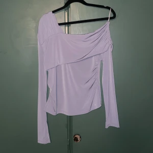 Grå topp från Gina Tricot - Supersnygg grå topp från Gina Tricot med lång ärm och en axelbandslös detalj på ena sidan. Toppen har draperad effekt och sitter asymmetriskt över axlarna, vilket ger en unik och trendig look. Perfekt för dig som vill sticka ut lite extra! Använd 1 gång.