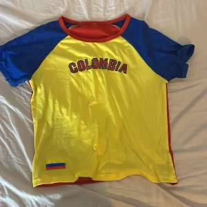 Colombia t-shirt i starka färger - Färgstark t-shirt med Colombia-tryck på bröstet. Framsidan är gul med blå ärmar och röd krage, baksidan är helröd. Liten Colombia-flagga nertill. Perfekt för dig som vill sticka ut och visa support. Skön och lätt i syntetmaterial. OBS!Köpt utomlands och liten i strl.