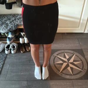 Svart tight kjol med röd och vit rand - Snygg svart kortkjol med tight passform och sportiga röda och vita ränder längs sidorna. Kjolen sitter högt i midjan och har en stilren, enkel design som ger en cool och modern vibe.