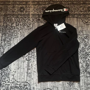 Svart zip-hoodie från Champion - Svart hoodie från Champion med dragkedja och huva. Klassisk logga på bröstet och stor logga på huvan. Mjuk bomullskänsla, fickor framtill och ribbade muddar. Perfekt för chill eller streetwear-stil.