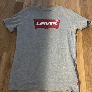 Grå Levi's t-shirt med röd logga - Snygg grå t-shirt från Levi's med klassisk röd logga framtill. T-shirten är i mjuk bomull och har en normal passform med korta ärmar. Perfekt till jeans eller shorts för en avslappnad look.