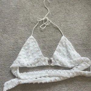 Vit topp med spets och knyt - Supersöt vit topp med spets och hålmönster. (Ej bikinimaterial). Triangelformade kupor, smala band att knyta i nacken och runt ryggen samt en liten  ring i mitten. 