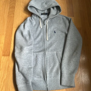 Grå hoodie från Polo Ralph Lauren - Snygg grå hoodie från Polo Ralph Lauren med dragkedja och klassisk broderad logga på bröstet. Tröjan har huva med vita snören och två fickor framtill. Perfekt för chill dagar och enkel att matcha med jeans eller joggers. OBS! Tröjan är lite nopprig här o där!!!?!! 
