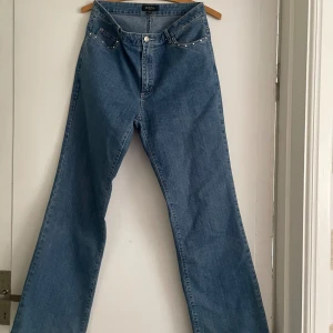 Blå raka jeans med nitar från G&FA - Snygga blå jeans från G&FA med bootcut-ben och hög midja. Jeansen har coola silverfärgade nitar längs fickorna fram och klassiska bakfickor. Perfekta för dig som gillar en retro vibe och vill sticka ut lite extra.