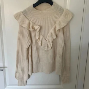 Beige stickad tröja med volang - Superfin beige stickad tröja med ribbad struktur och volangdetaljer över axlar och bröst. Hög hals och långa ärmar ger en mysig och trendig look. 