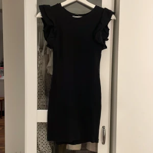 Svart volangklänning från Zara - Supersnygg svart kortklänning från Zara med volangdetaljer på axlarna. Klänningen är ribbad och har en tight passform som framhäver figuren. Perfekt för dig som gillar stilrena och trendiga plagg med det lilla extra.